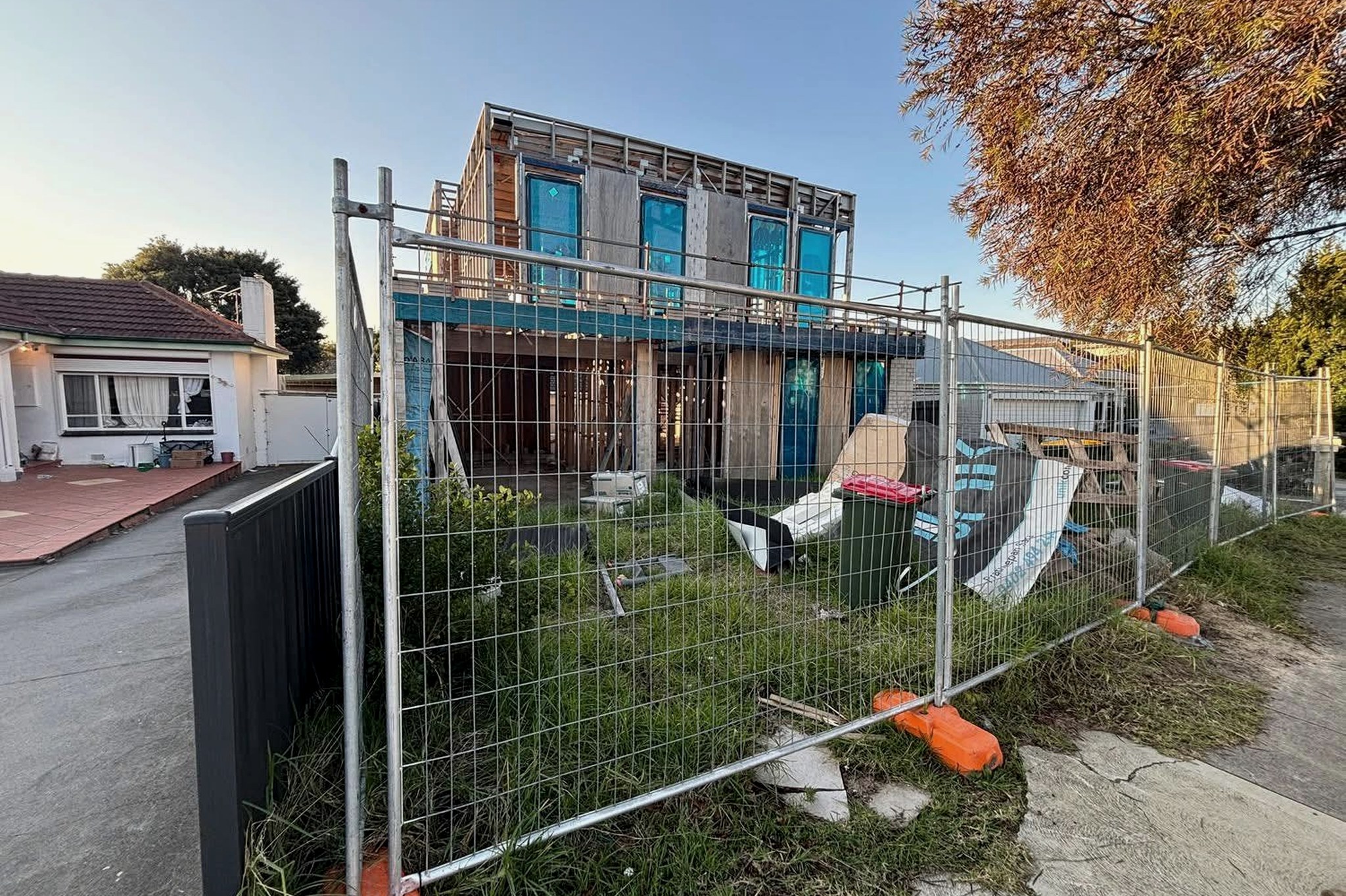A partially-built residential construction site behind temporary fencing, overgrown grass and construction debris visible.