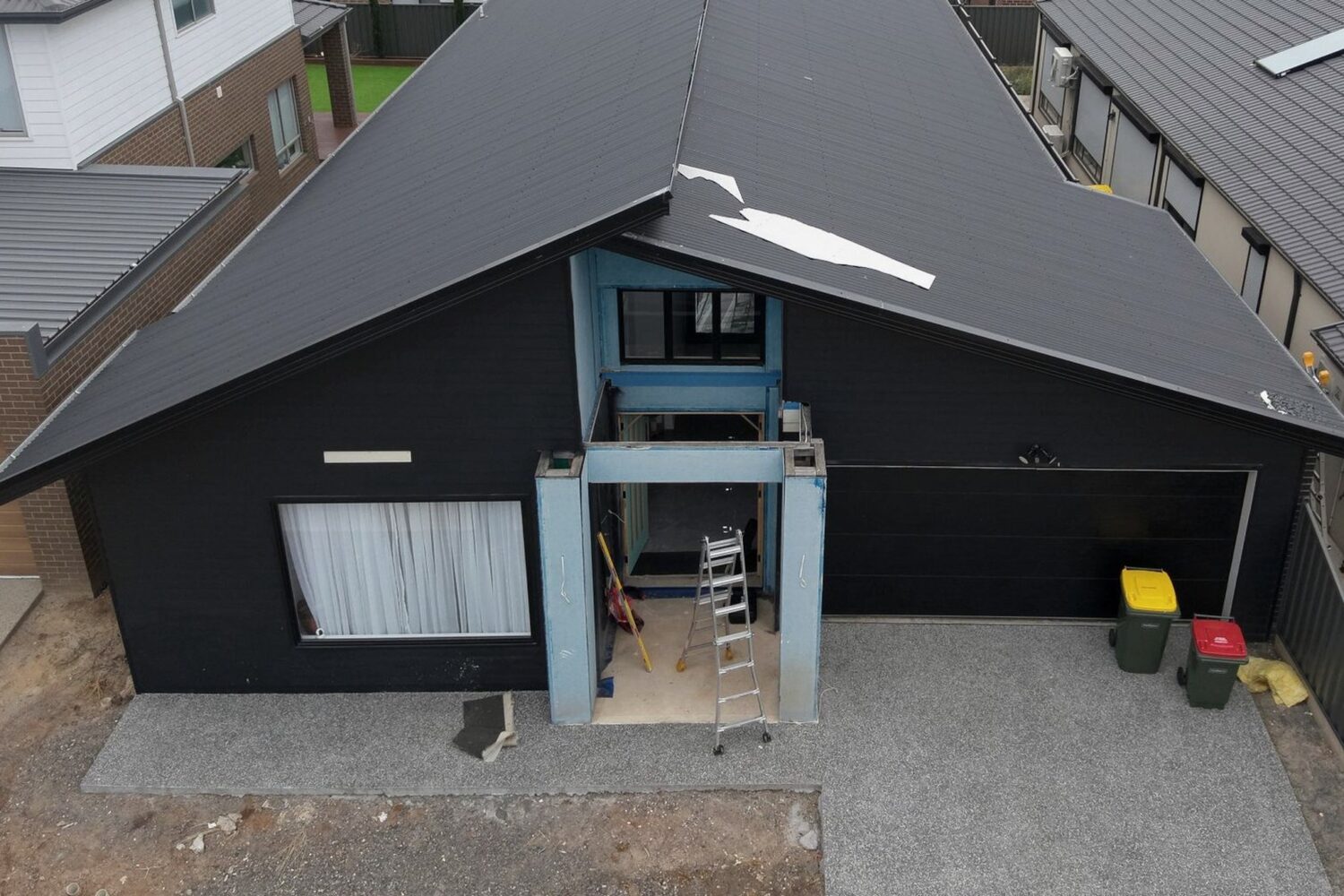 Aerial view of a single-storey home, occupied but unfinished: a ladder in the open entry, protective film still on the entry columns and bare earth where landscaping should be.
