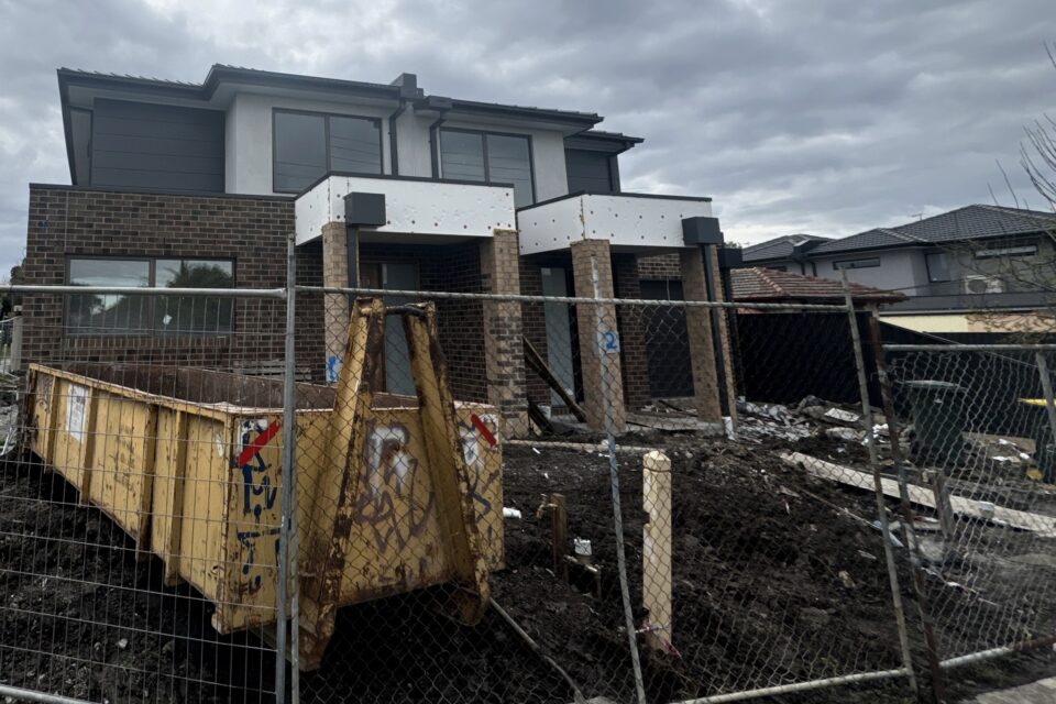 A two storey brick home still under construction behind temporary site fencing, with a skip bin, bare earth and exposed insulation board at the balcony fascia