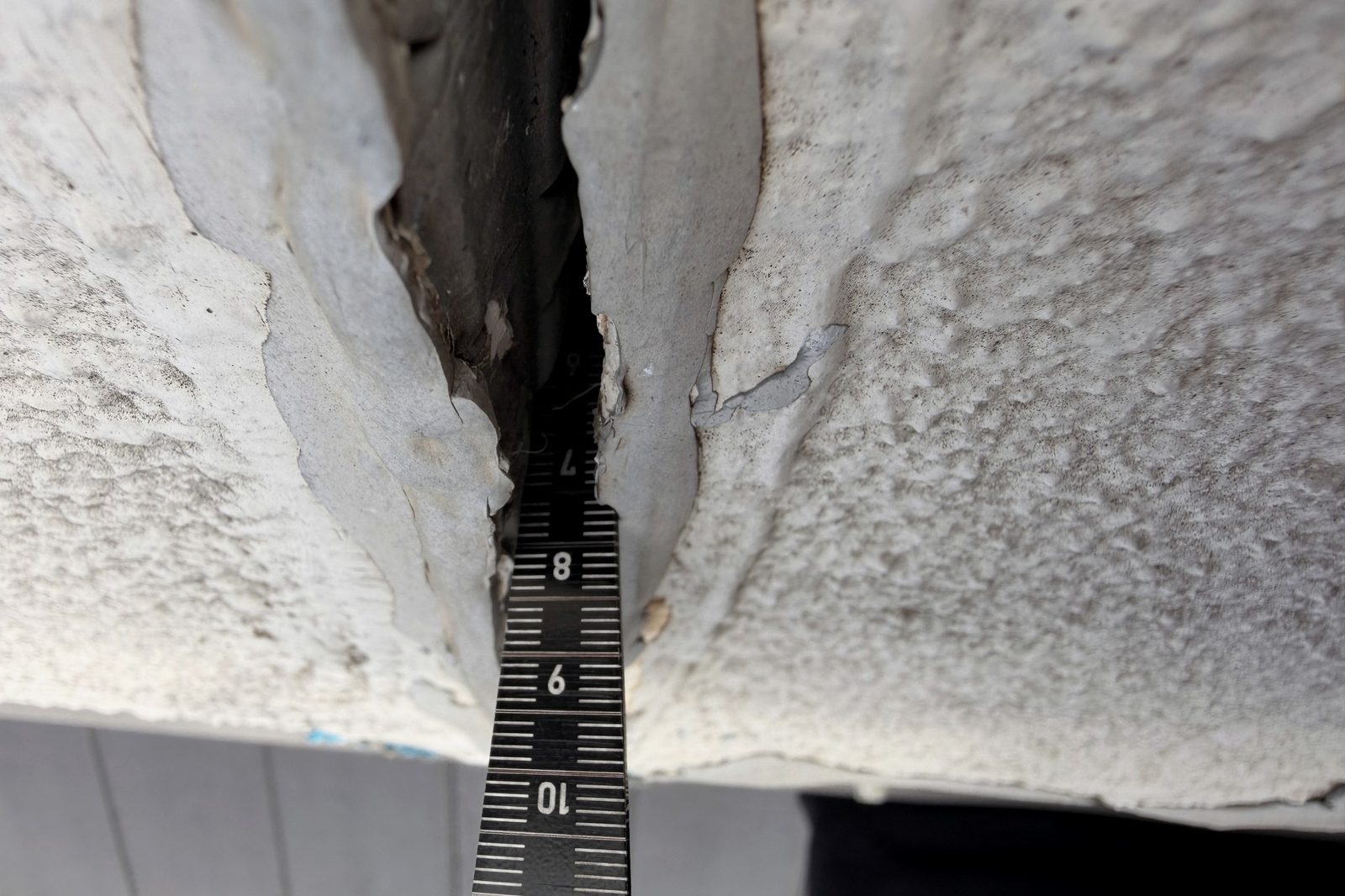 A steel rule held against a separation at a ceiling junction, recording the gap in millimetres