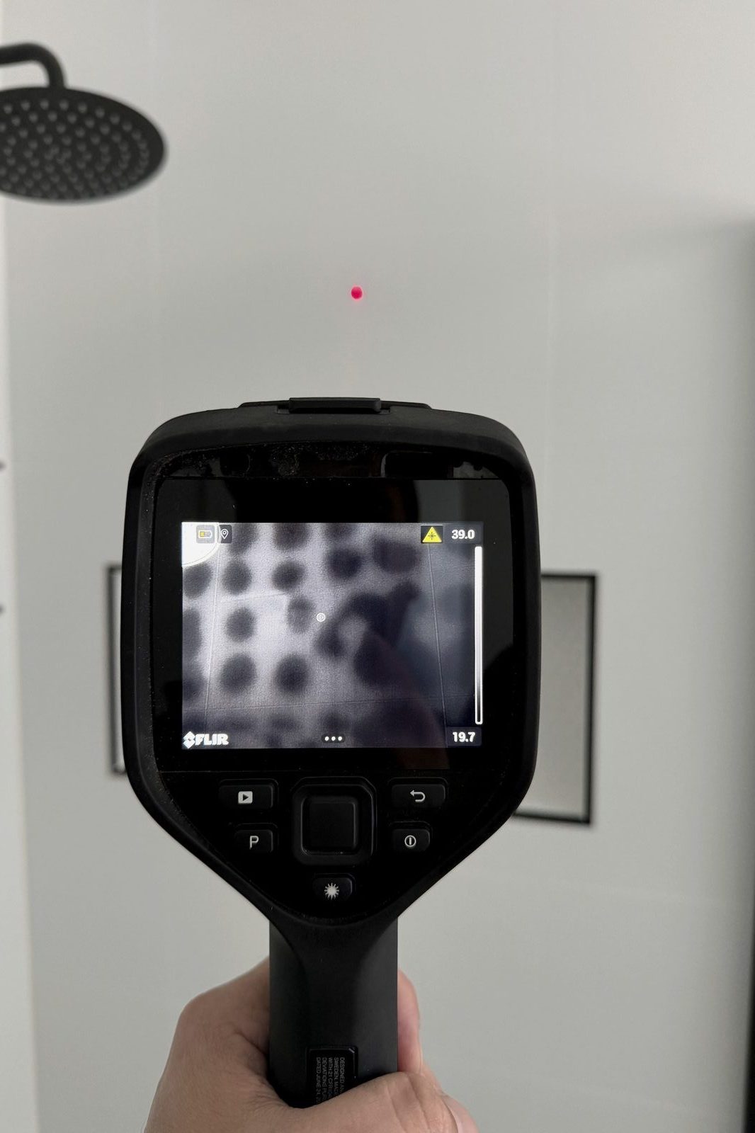 A thermal imaging camera held against a wall during an inspection, its screen showing cooler areas