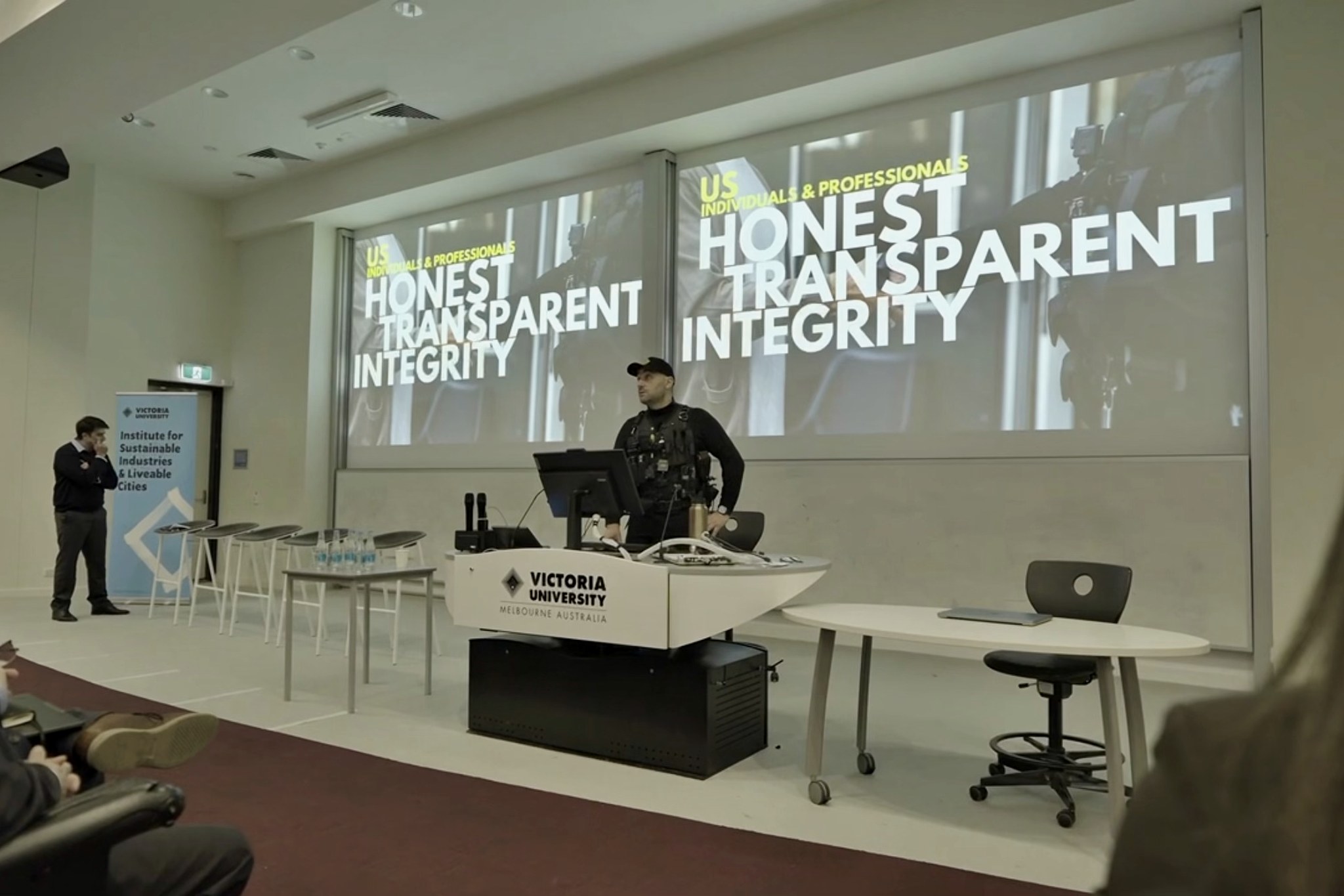Zeher Khalil speaking at a Victoria University lectern with his presentation on screen behind him