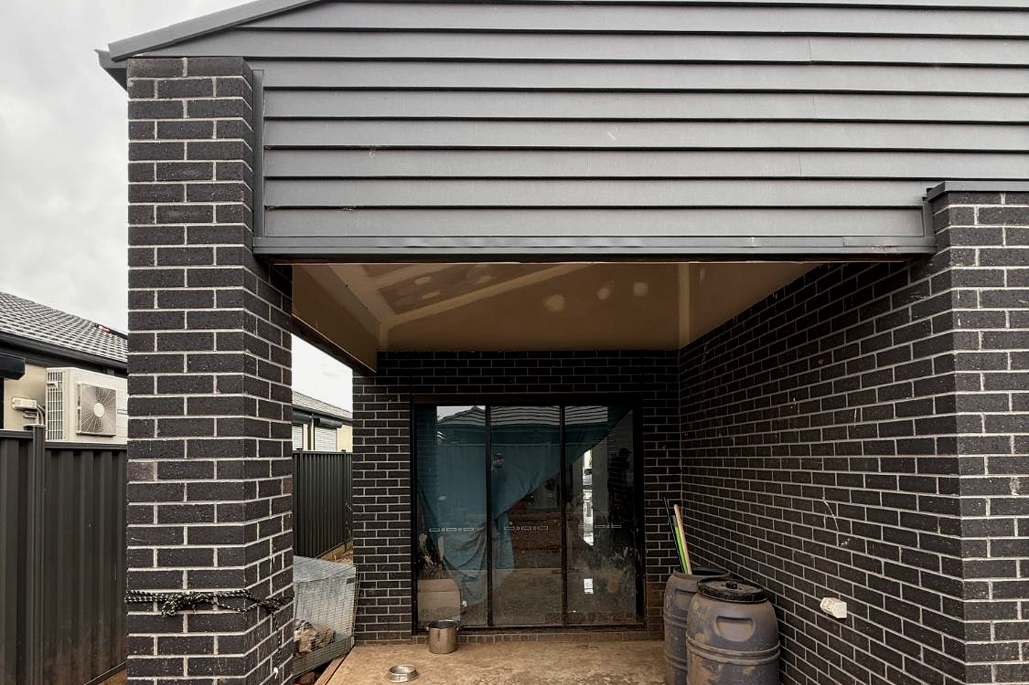 Rear alfresco of the same home: unsealed concrete, bare earth where paving and landscaping should be, builder plastic still on the glazing, and dog bowls and a lead showing the home is lived in.