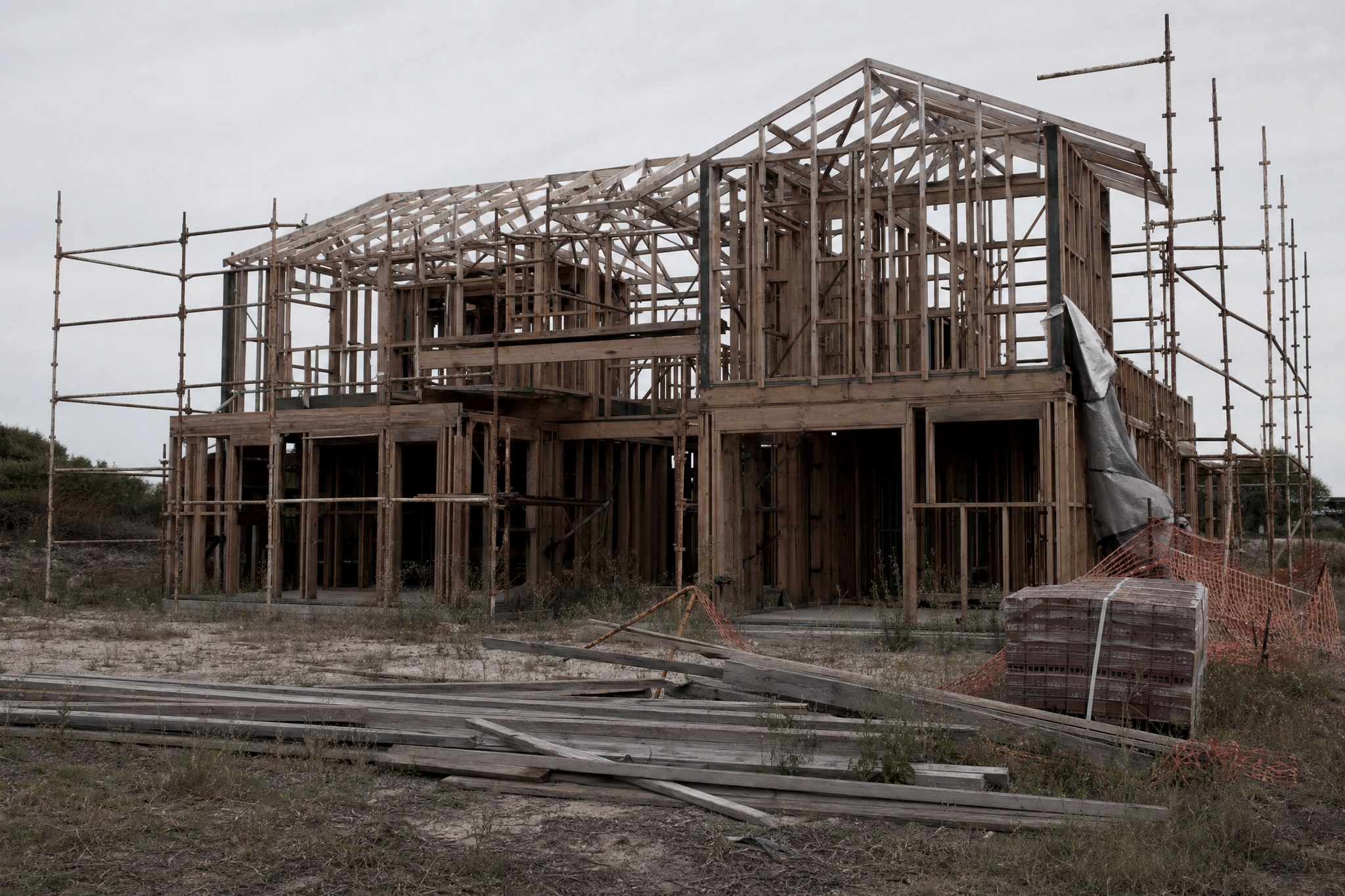 A deserted two-storey house at frame stage, timber frames exposed with no cladding, weathered framing timber and a strapped pallet of bricks lying in weeds, scaffolding still standing.