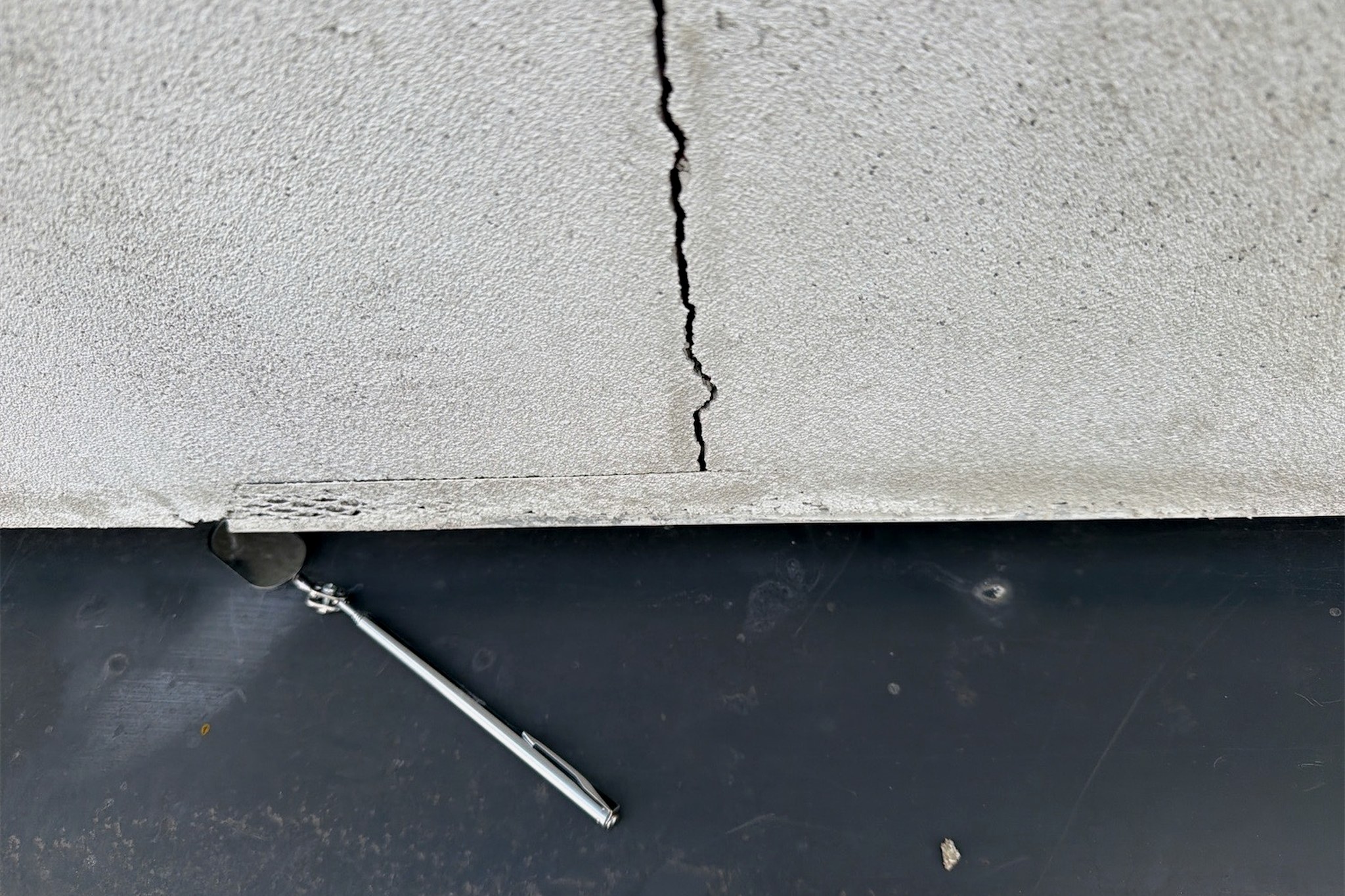 A crack running through a rendered element and opening wider towards the edge, with an inspection probe placed alongside for scale