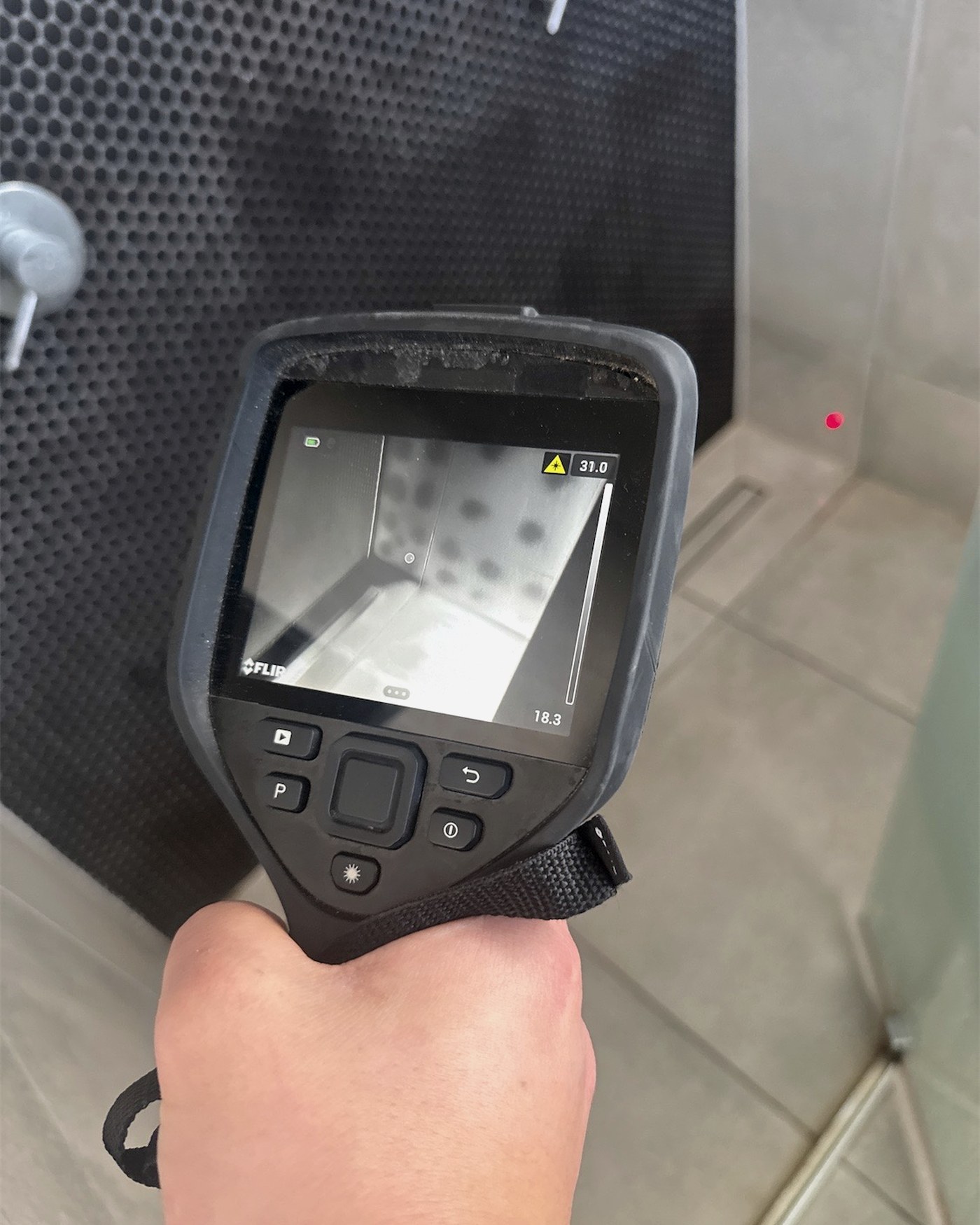 A thermal imaging camera held up to a tiled shower wall, its screen showing a regular pattern of cooler circular patches behind the tiles
